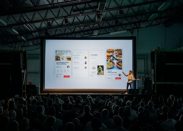 Man in yellow shirt points to large screen showing slides in front of large, dimmed audience